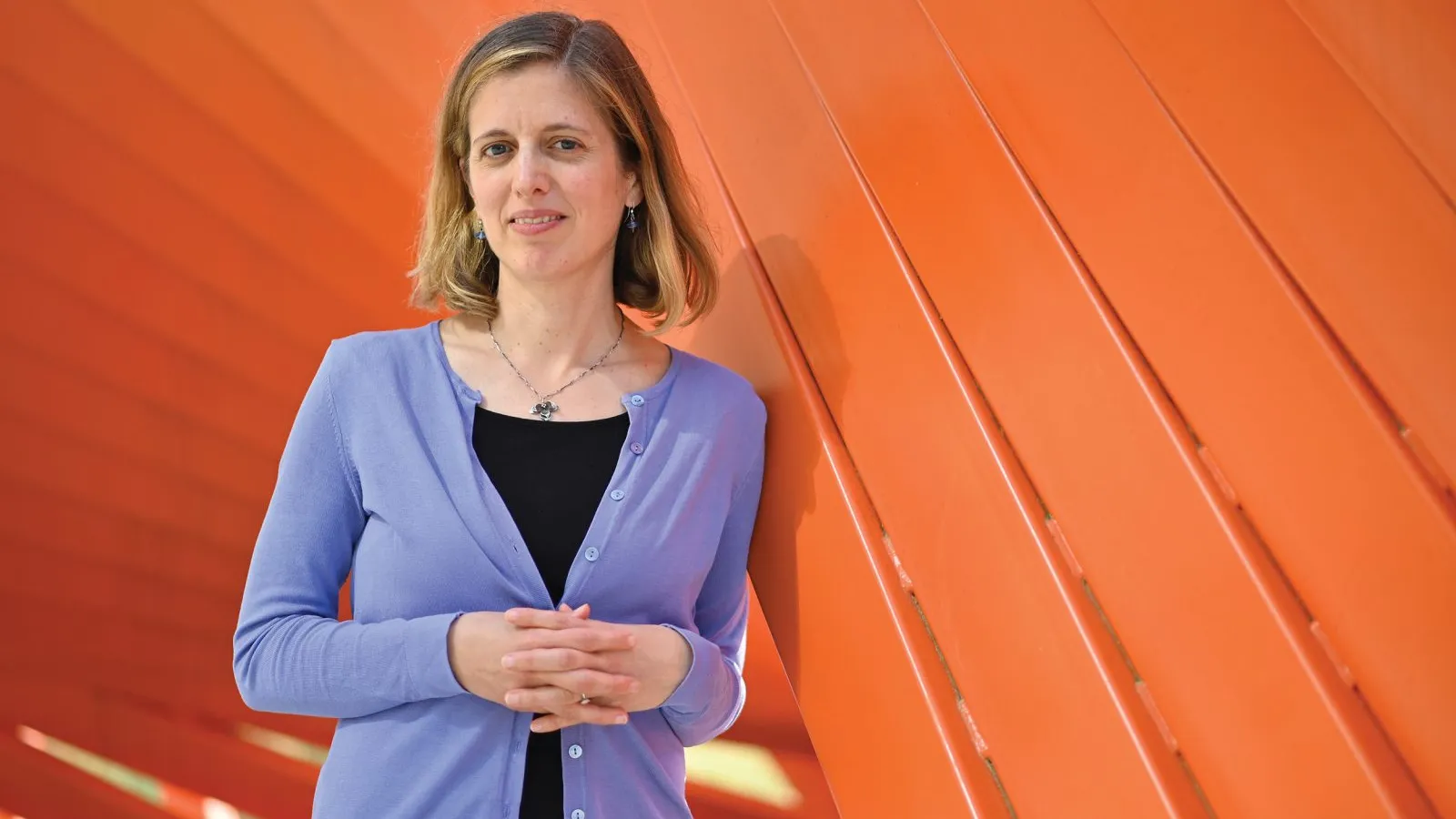 A blonde woman in a purple cardigan leaning against a wall.
