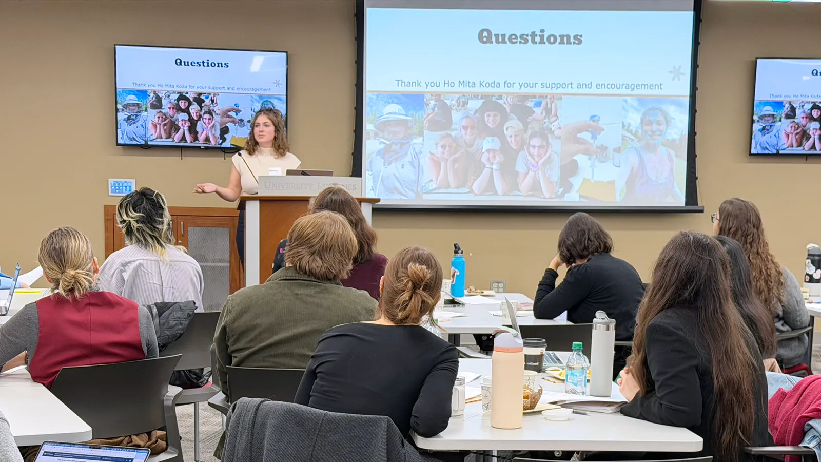 Student standing at podium beside projection screen in front of seated audience.