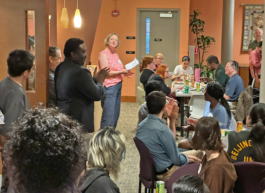 Diverse group of people gathered in an open space of an office suite, some standing and some sitting, listening to a speaker.