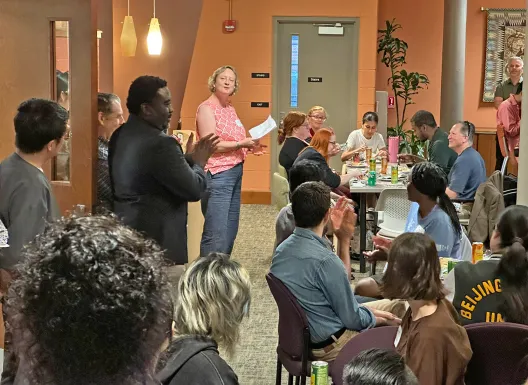 Diverse group of people gathered in an open space of an office suite, some standing and some sitting, listening to a speaker.