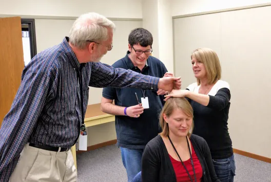 Woman sitting in chair as two people hold up her hair and another is ready to cut it
